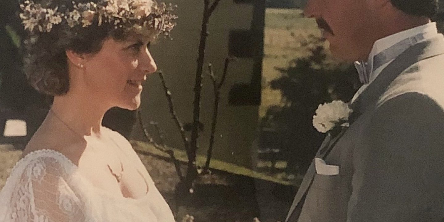 Robyn on her wedding day with short brown hair in white wedding dress holding hands with Grant who has dark hair and a moustache wearing a grey suit.