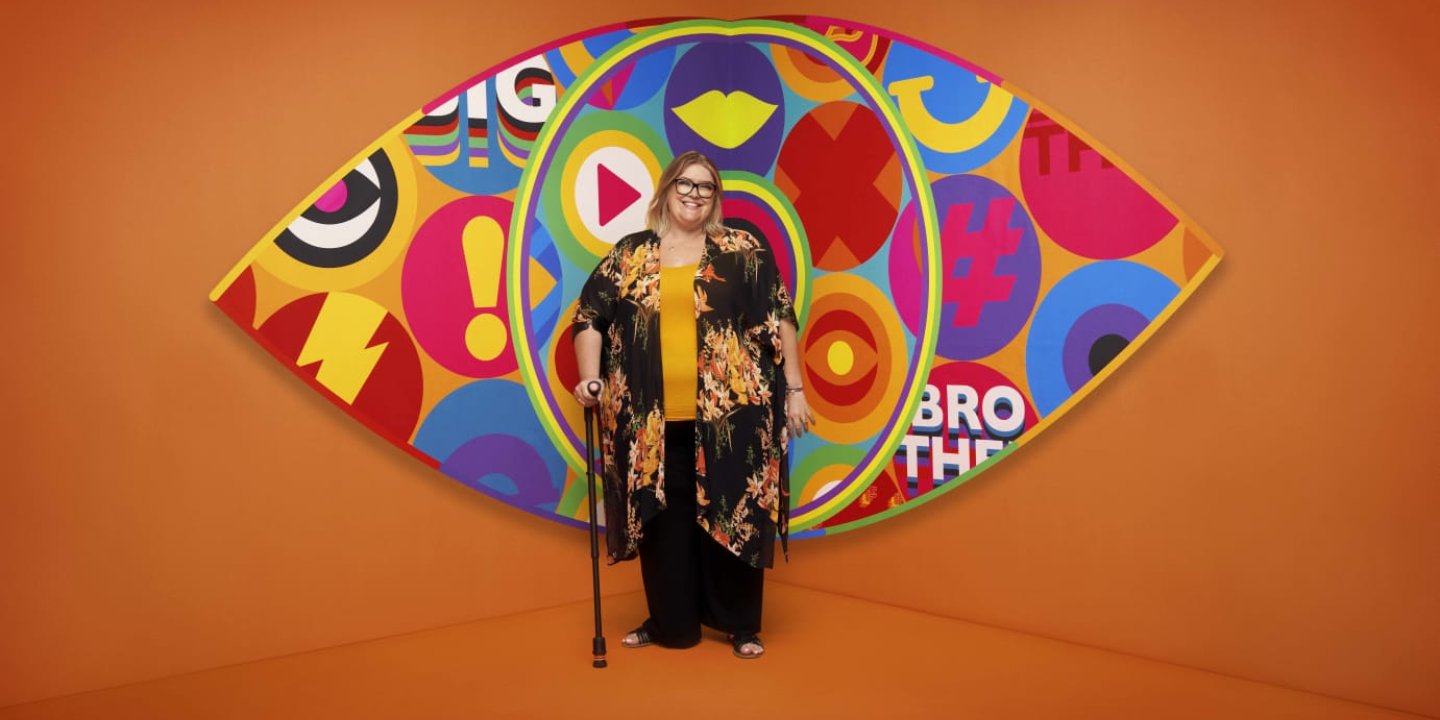Kerry stood with a stick wearing a colourful outfit in front of the Big Brother eye symbol and an orange background