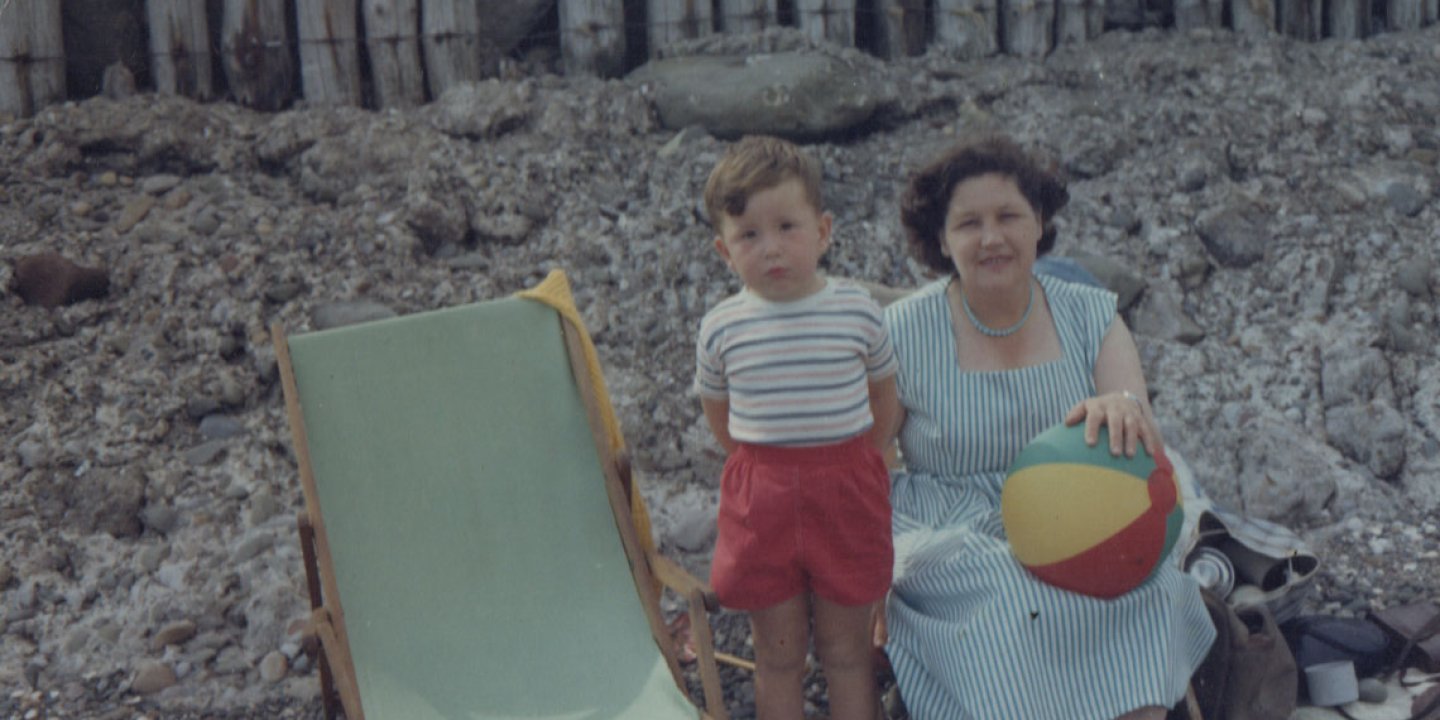 Dave Myers as a child on the beach with his mother. His mother is sat on a chair holding a beach ball and Dave is stood next to her in red shorts and a stripy t shirt.
