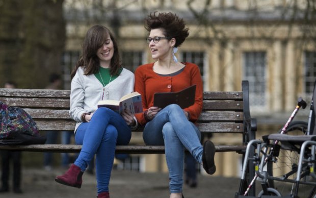 Photo shows two young women sitting on a bench, reading. 