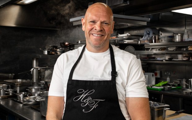 Tom is standing in his restaurant's kitchen wearing a white t-shirt and a black apron. He's smiling at the camera with his arms down by his sides.