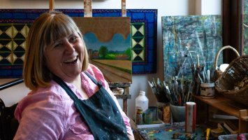 Jenny sits surrounded by art materials, wearing a paint-splattered apron and with a big smile