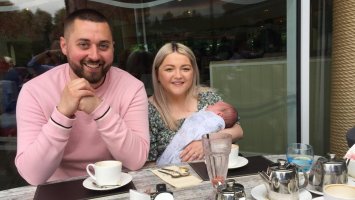 Ryan and partner Emma, holding their baby, smiling sat at a garden table
