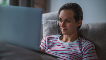 Woman, propped up on pillows on a sofa looks at a laptop