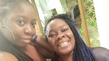 Elisha to the left wears hoop earing and has long braided hair, her suntie to the right smiles at the camera with long braided har that's blue and black. 