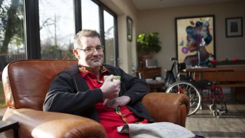 Mark sits in a brown leather armchair holding a mug. Behind him are windowed doors looking into a garden. Mark's wheelchair is in the background. 