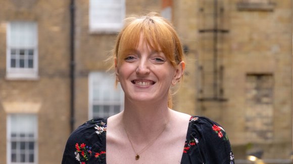 A headshot of Charlotte Nichol MP, a woman with red hair with a fringe in a pony tail, smiling.