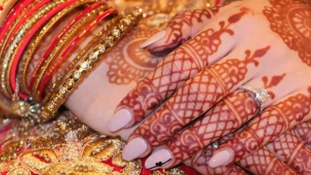 Clasped manicured hands with mehndi decoration rest on a rich red and gold background