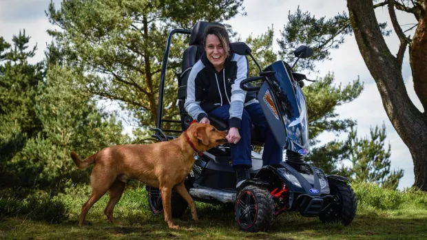 The photo shows a woman sitting in a TGA mobility scooter. She's smiling and playing with her dog. 