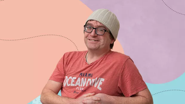White man wearing glasses and a knitted beanie hat grins to camera