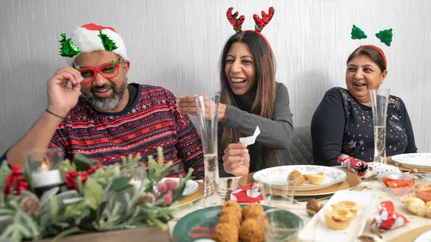 A family sit around a table laden with food and Christmas crackers. They wear funny festive hats and festive jumpers and are laughing at a joke from a cracker