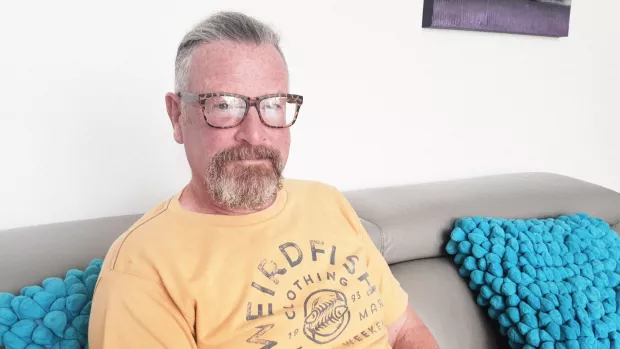 A man wearing a yellow t-shirt and glasses sits on a sofa and looks to camera