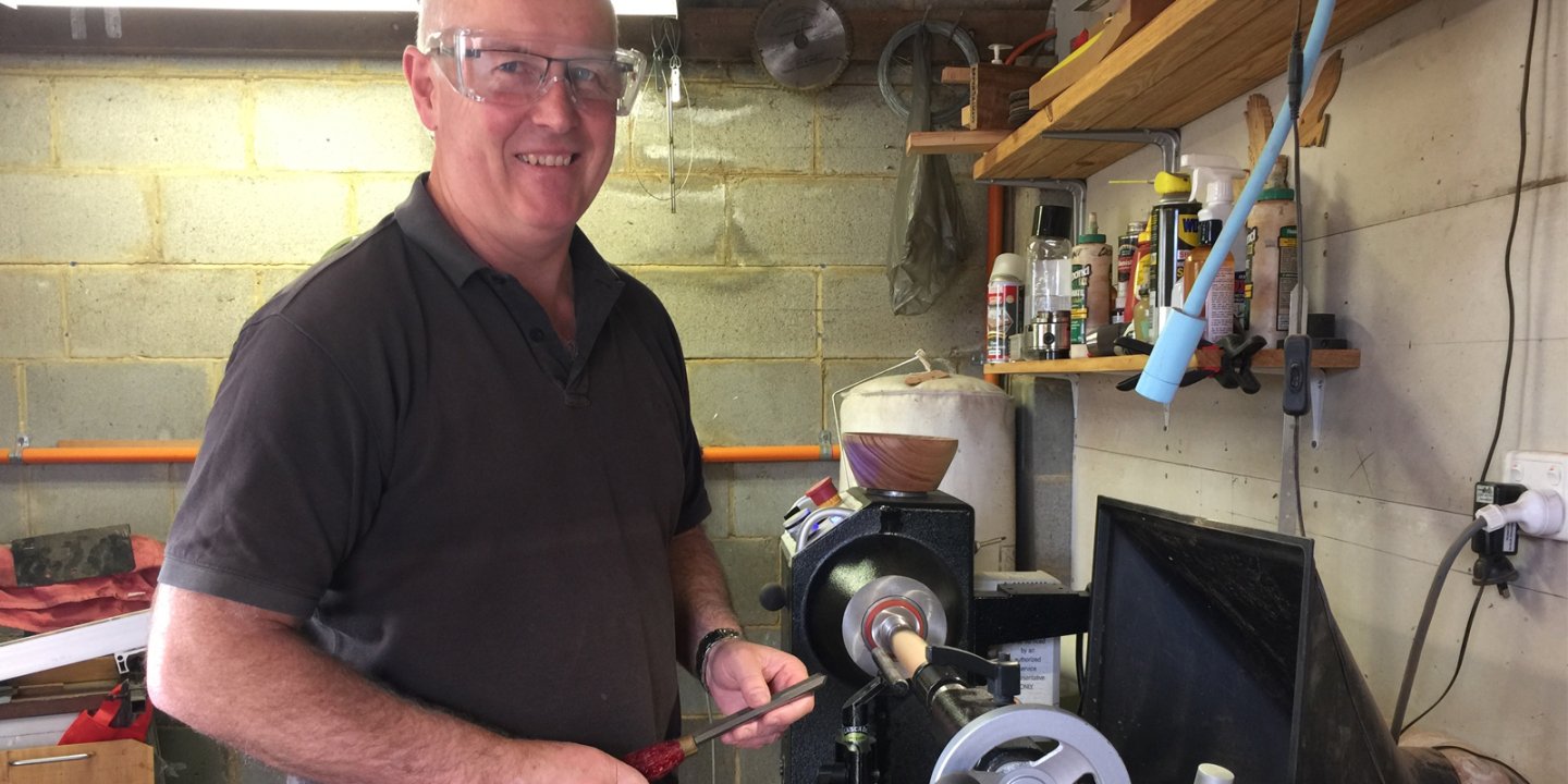 Grant wearing a plaid shirt smiling with a wooden vase he's made on his lathe