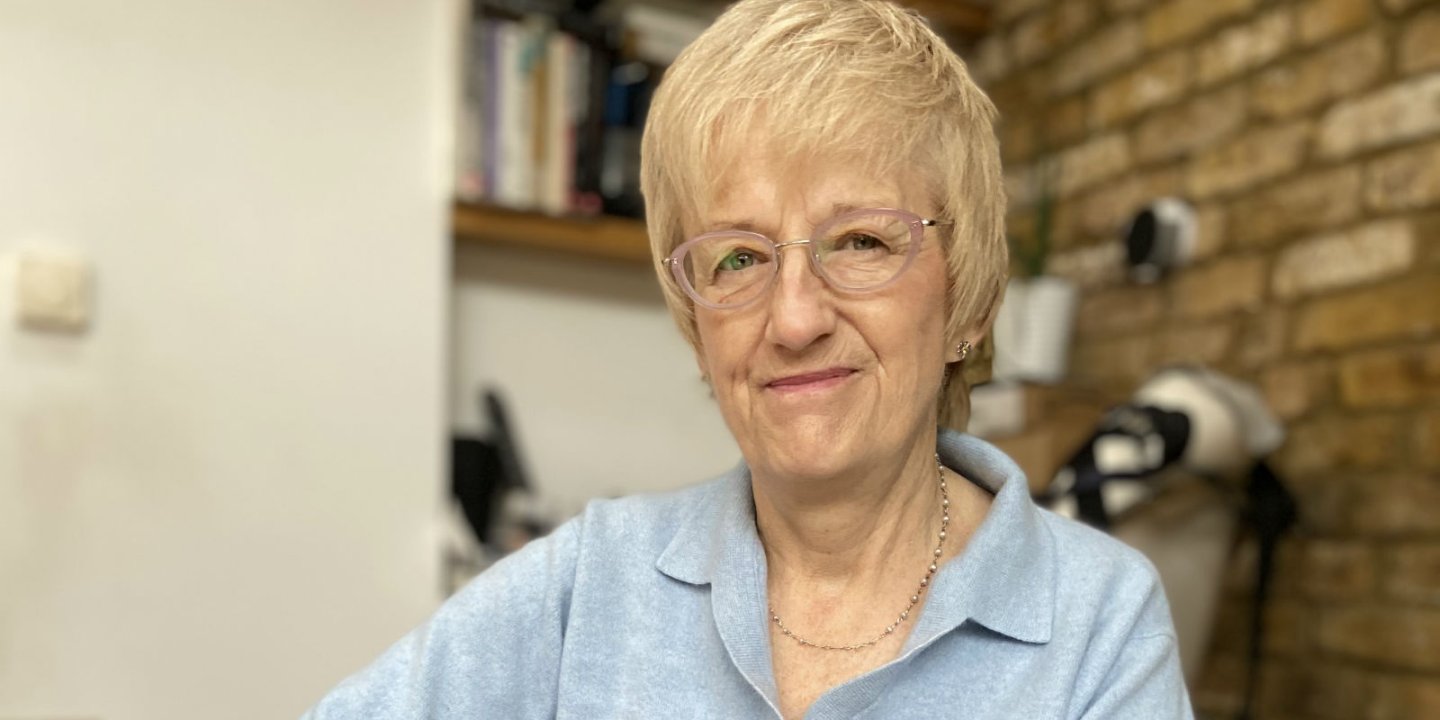 Christine has short blonde hair and glasses. She sits in her house.