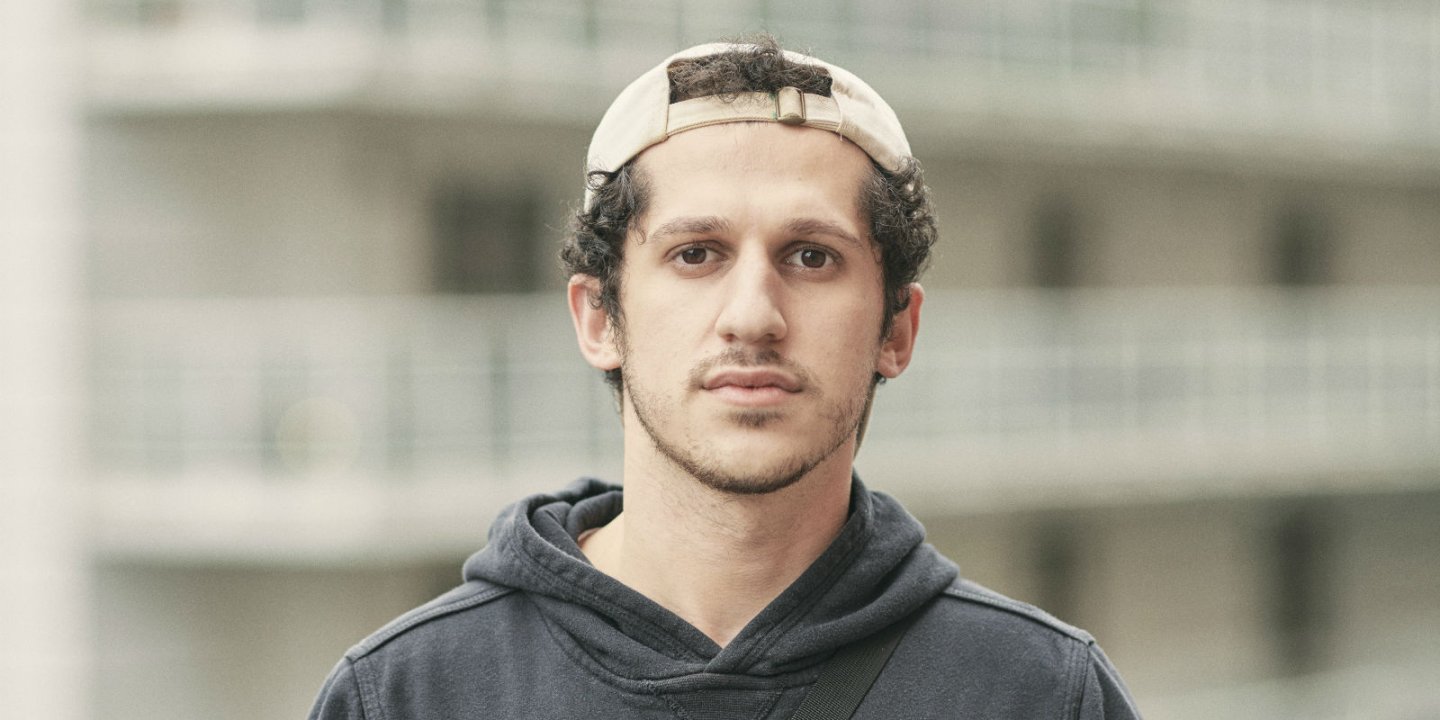 Ayad wears a sweatshirt and baseball cap, in front of a block of flats.