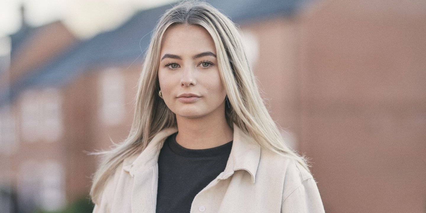 Head and shoulder portrait of Ellie who has long blond highlighted hair looking seriously to camera with brick houses blurred outbehind her.