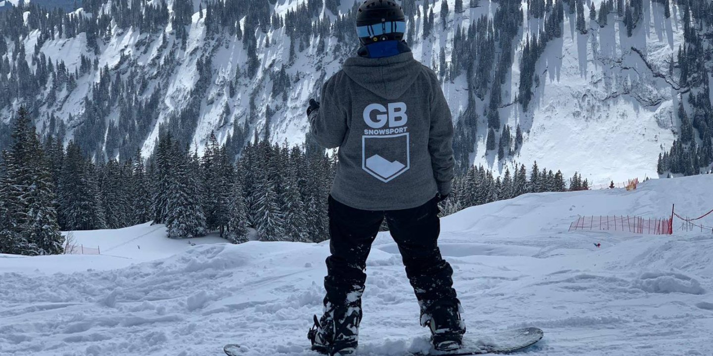 Nina stands on her snowboard facing away from the camera to a view of a snowy, rocky mountain and fir trees.