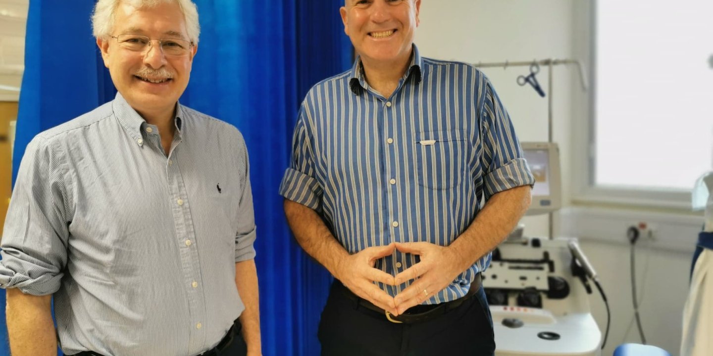 Two men, Basil (left) and John (right) wearing shirts and black trousers stand smiling with their backs against a blue curtain and hospital equipment in the background