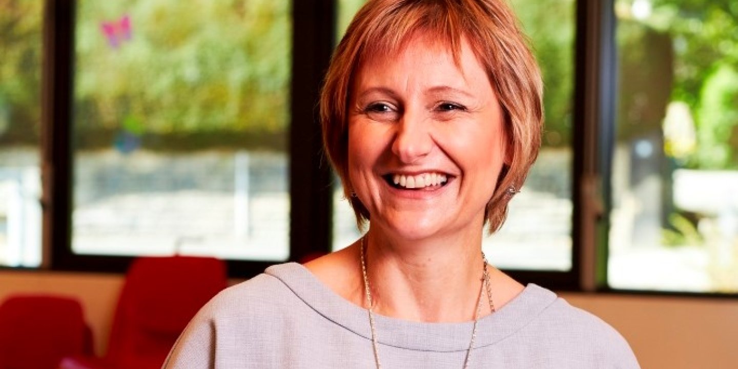 Image shows a headshot of a smiling woman. She has short hair and it wearing a light coloured top, and the background is blurred greenery through a window. 