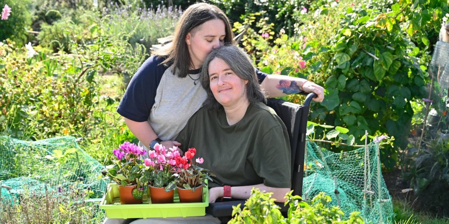 Sarah stands by her wife Karine, who is using a wheelchair. Sarah kisses her on the top of her head.