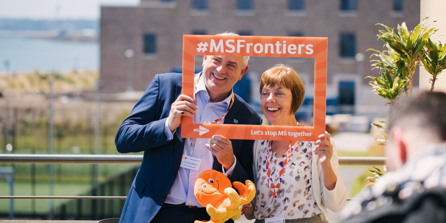 Two researchers holding up an orange frame with #MSFrontiers on it.