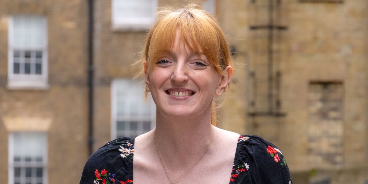 A headshot of Charlotte Nichol MP, a woman with red hair with a fringe in a pony tail, smiling.