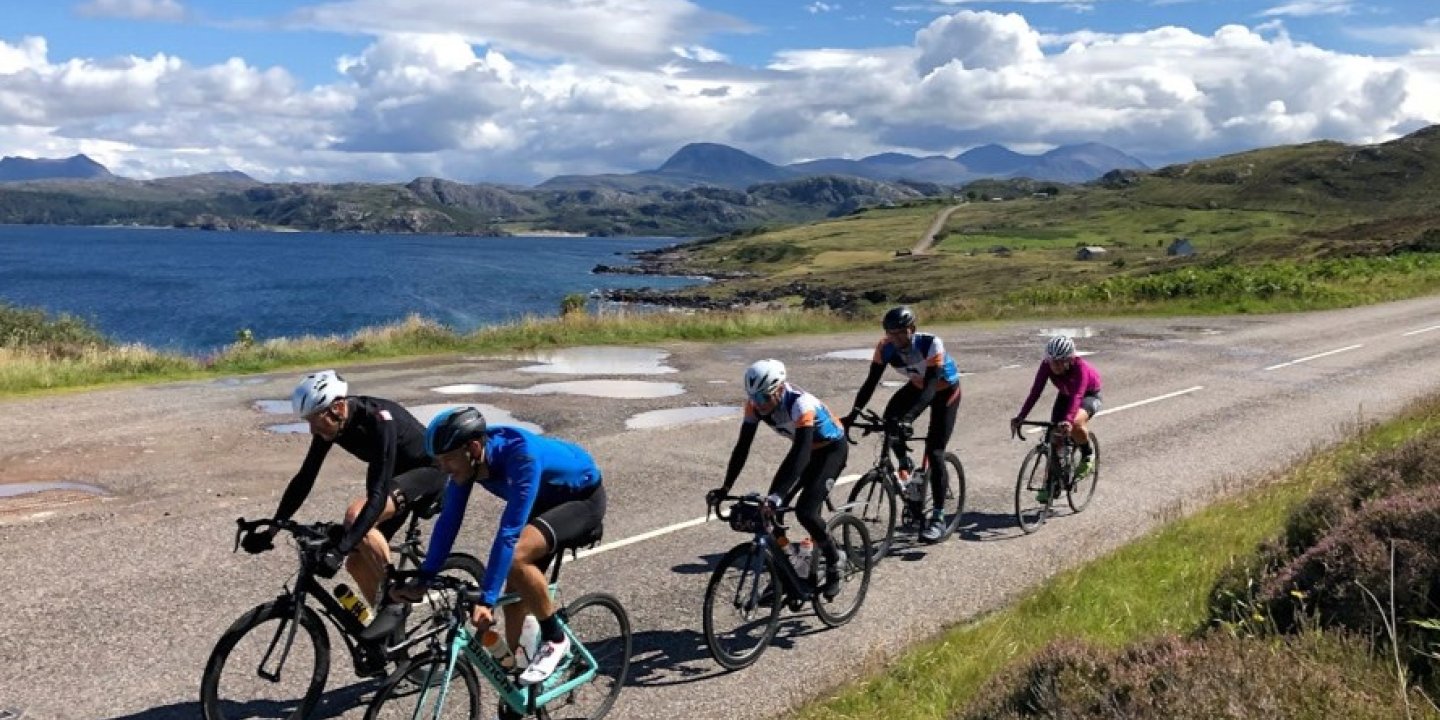 A group of five cyclists riding along a road in front of a late and hills