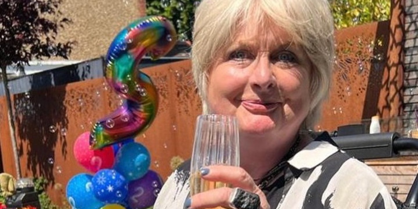 Jane sits in the sun holding a champagne flute, with a pile of colourful balloons in the background.