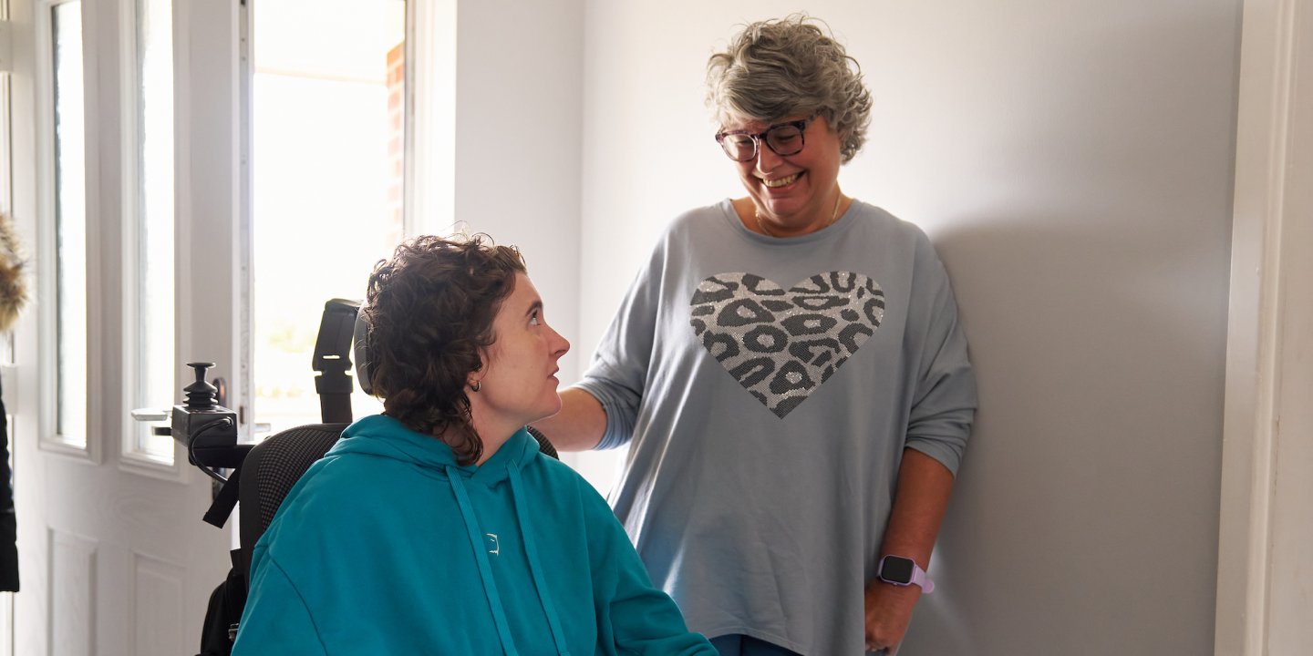 Jasmine and her mum are in the hallways of Jasmine's house with the front door behind them. They're smiling at each other and her mum rests her hand on the back of Jasmine's wheelchair.