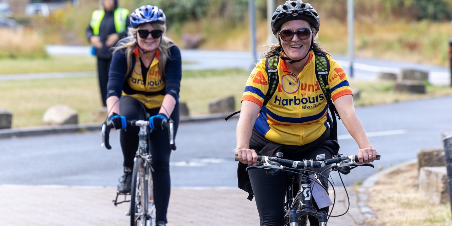 Two people cycling wearing yellow 'Round the Harbours' jerseys