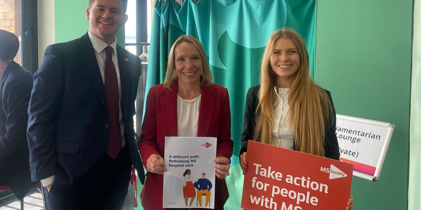 Three people smiling at the camera, one holding a sign saying Take action for people with MS