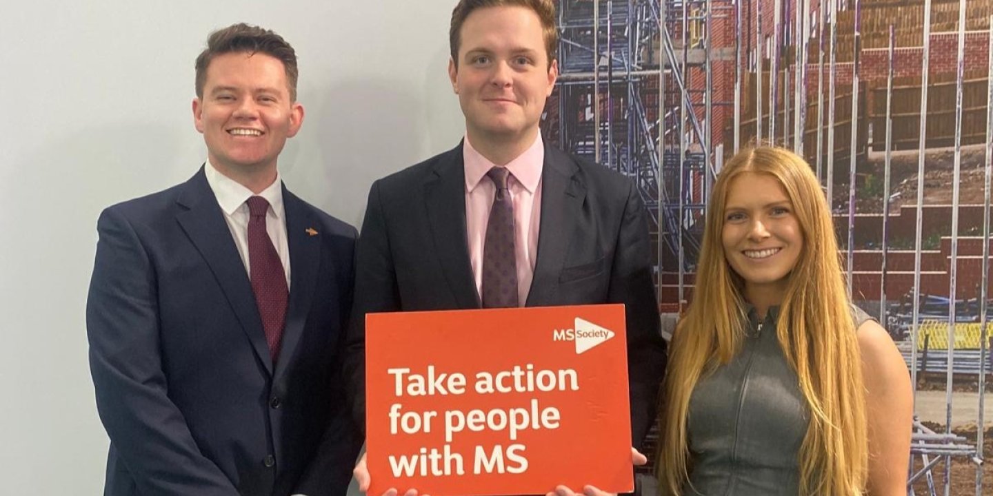 Three people smiling at the camera, one holding a sign saying Take action for people with MS