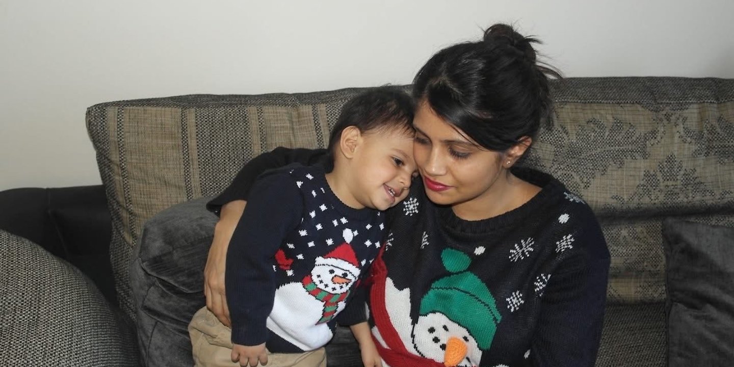 Niti and her son, Nico are sat on a sofa hugging, they both wear Christmas jumpers with snowmen on.