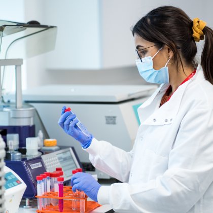 In a lab, a researcher wears a face mask, blue gloves, and a lab coat while checking the vials.