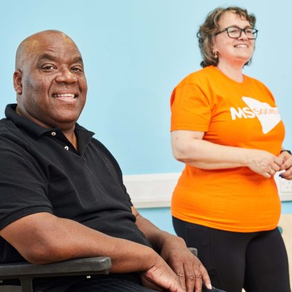 A man in a wheelchair smiles at the camera, with a person in an MS Society t-shirt in the background