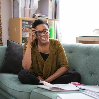 A woman sits cross legged on a green sofa with papers spread out in front of her