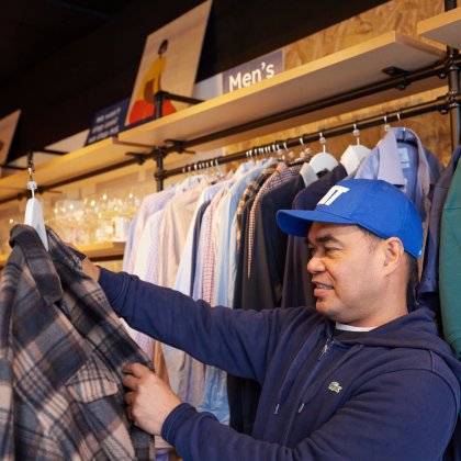 A man in a blue hoodie and baseball cap looks at a checked shirt from a rack in a charity shop
