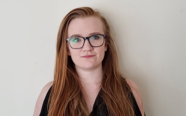 Fiona stands against a white wall. She has long red hair and is wearing glasses.