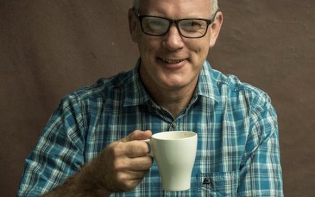 A man in a green checked shirt is smiling and holding up a white mug