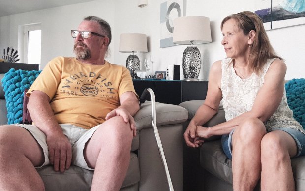 A man in shorts and a yellow t-shirt sits on a stone coloured sofa with a walking aid propped up against it, a woman in jean shorts and sleeveless top sits next to him. Both stare to the left as if out of a window. 