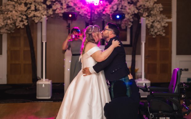 A wedding couple embrace for their firs dance, she is wearing a white wedding dress and he is in a kilt, behind him is his wheelchair from which he's just stood up