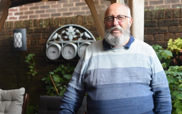 Mike has a thick white beard and wears glasses and a jumper with thick vertical stripes going from light to dark blue as they descend. Behind mike is a garden wall and some plants.