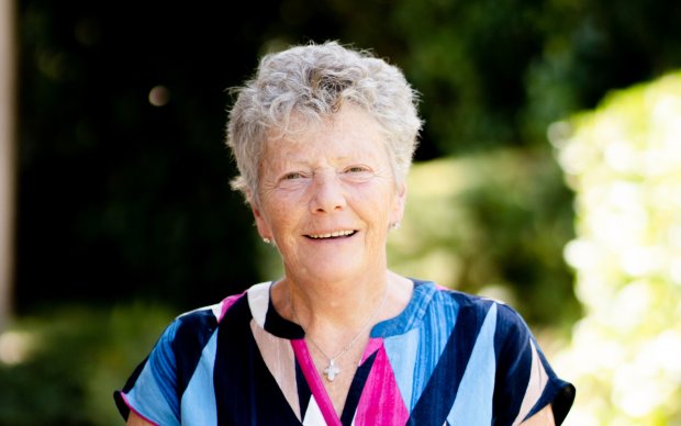 Siobhan has short wavy light grey hair and wears a colourful short sleeved top in pink and blue, behind them is the dappled light of of an out of focus summer garden.