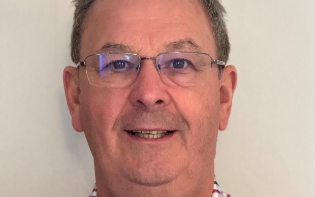 Hew has short dark grey hair, wears metal framed glasses and a blue and red plaid shirt that's open at the collar.