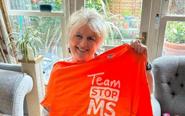 Jane holds up a bright orange 'Team Stop MS' t-shirt. Her dog sits in the foreground.