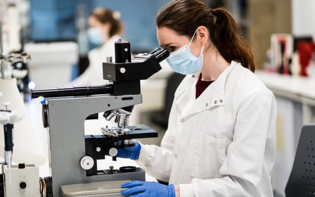 A researcher wearing a white lab coat, blue gloves and a blue face mask looking down a microscope
