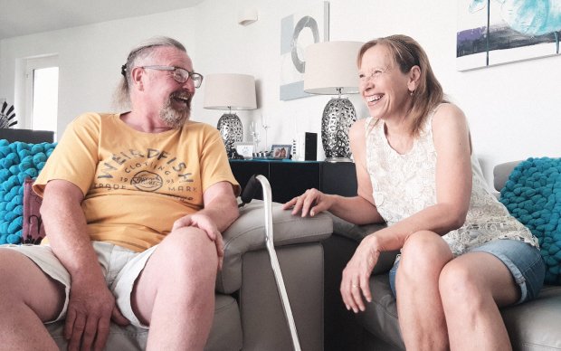 Ian wears a yellow t-shirt and shorts, he's sat on a sofa laughing and chatting