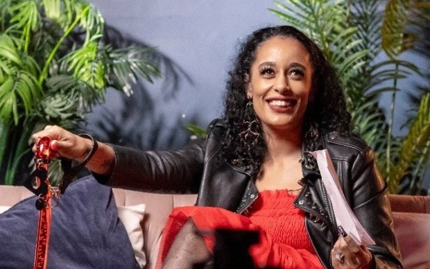 Roxy say on a sofa, smiling off camera. Wearing a red dress and black leather jacket and holding a red walking stick.
