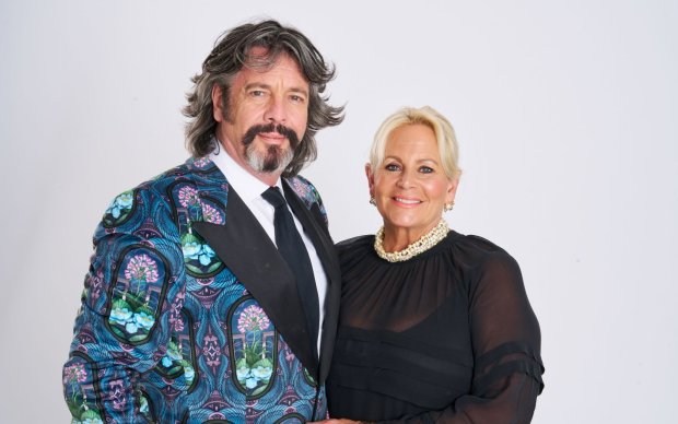 Professional portrait of Laurence and Jackie Llewelyn-Bowen against a white background. Laurence wears a turquoise patterned suit and Jackie wears a black dress and pearls.