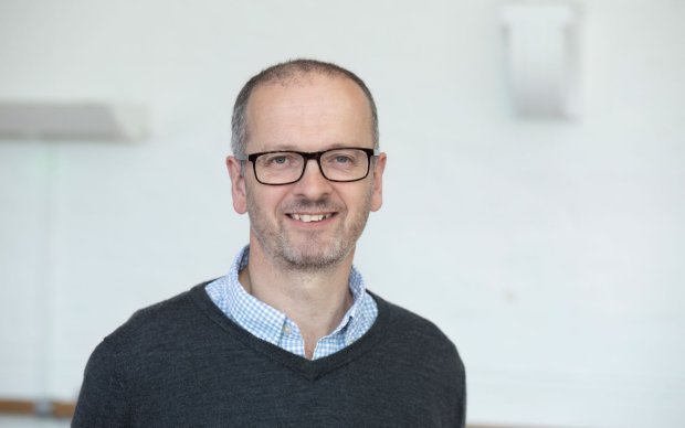 Headshot of Nick, who wears glasses and a shirt and jumper, in front of a white background